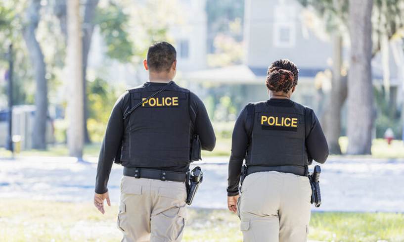Two uniformed police officers walk away from the camera. They wear black vests with the word "POLICE" in yellow across the back, and khaki pants.